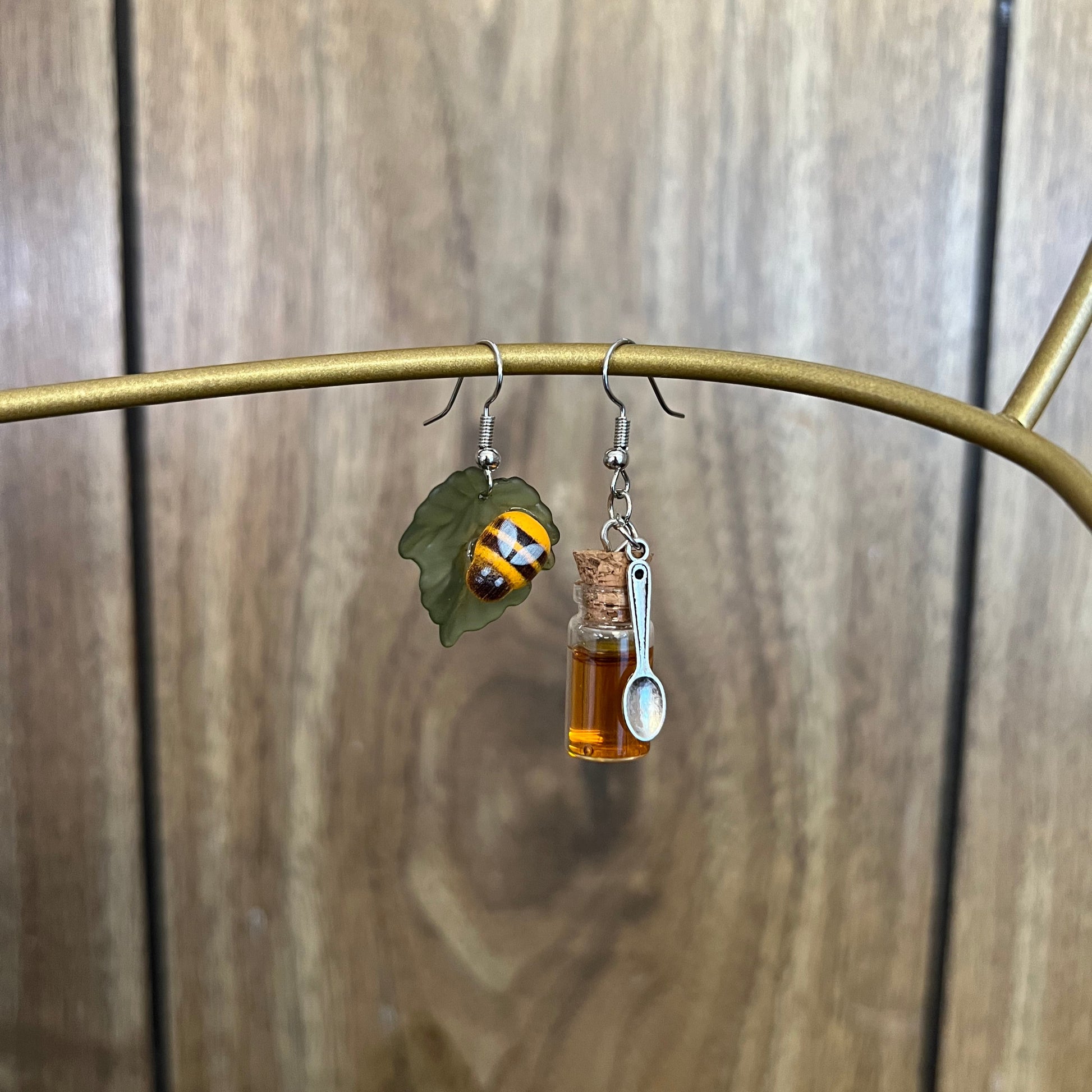 A pair of earrings featuring a bee and a honey jar design, displayed against a wooden background.