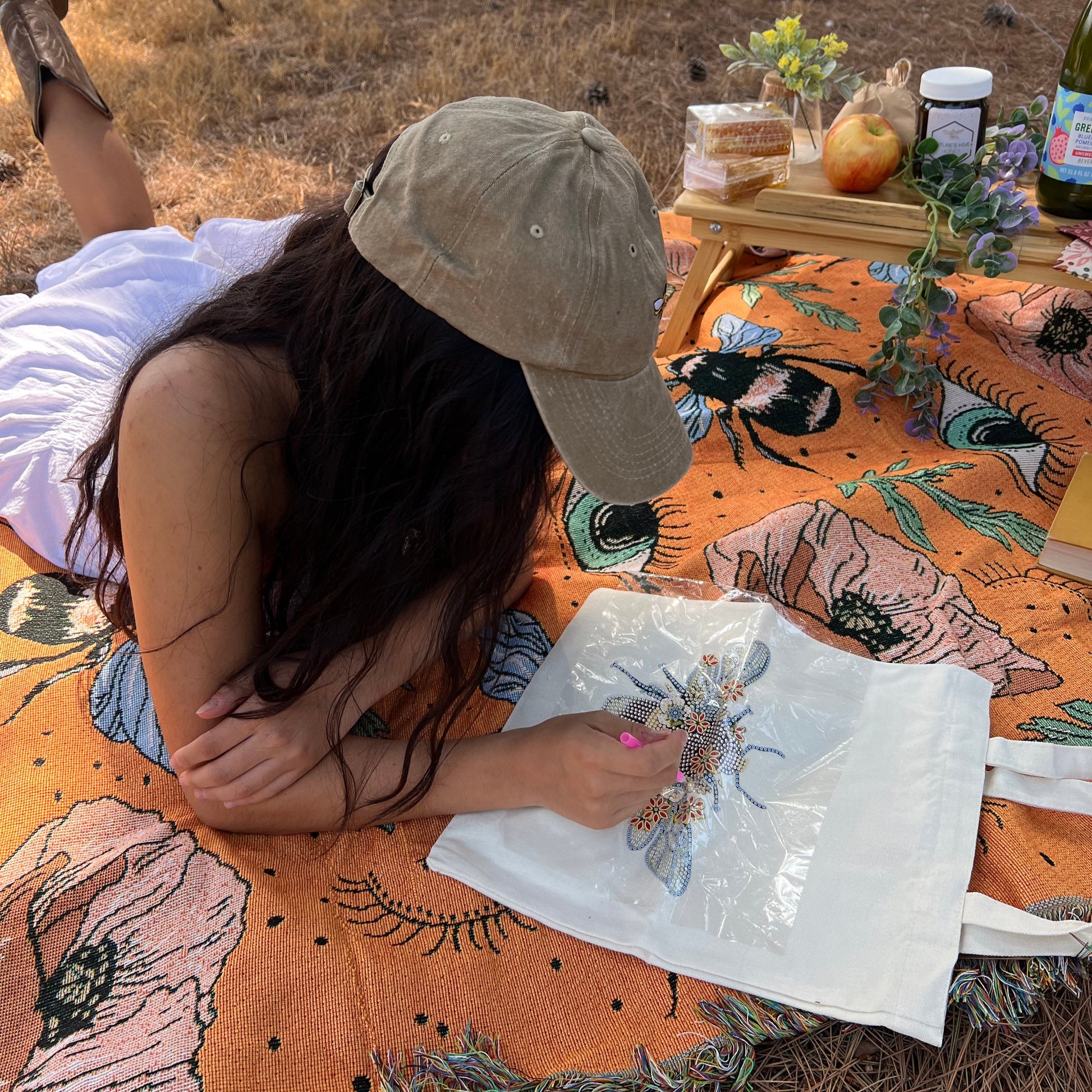 A person is sitting outdoors, drawing or coloring a design on a tote bag that features a blank canvas and is adorned with colorful rhinestones.