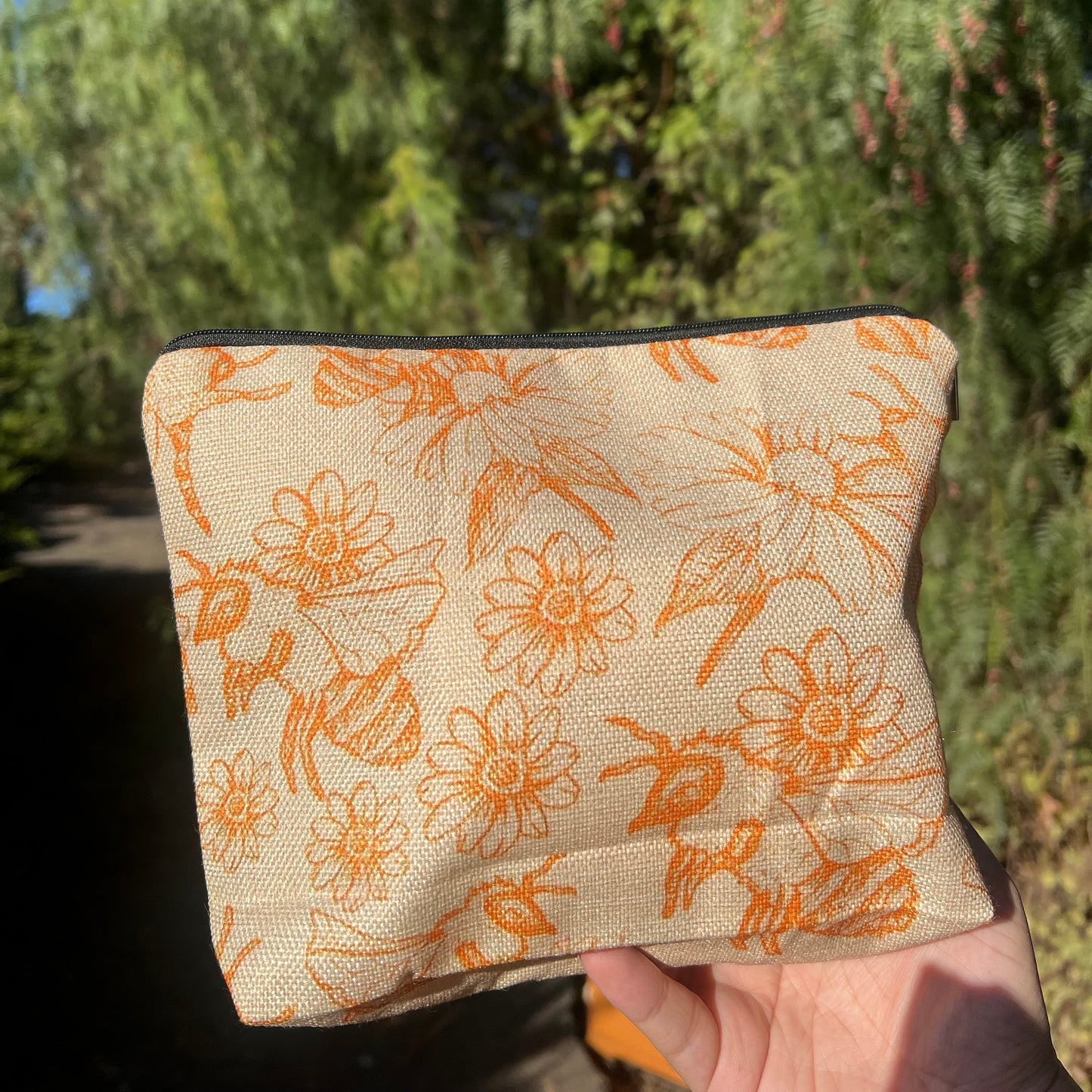 A beige cosmetics bag with orange floral patterns, including daisies and bees, held in a person's hand.