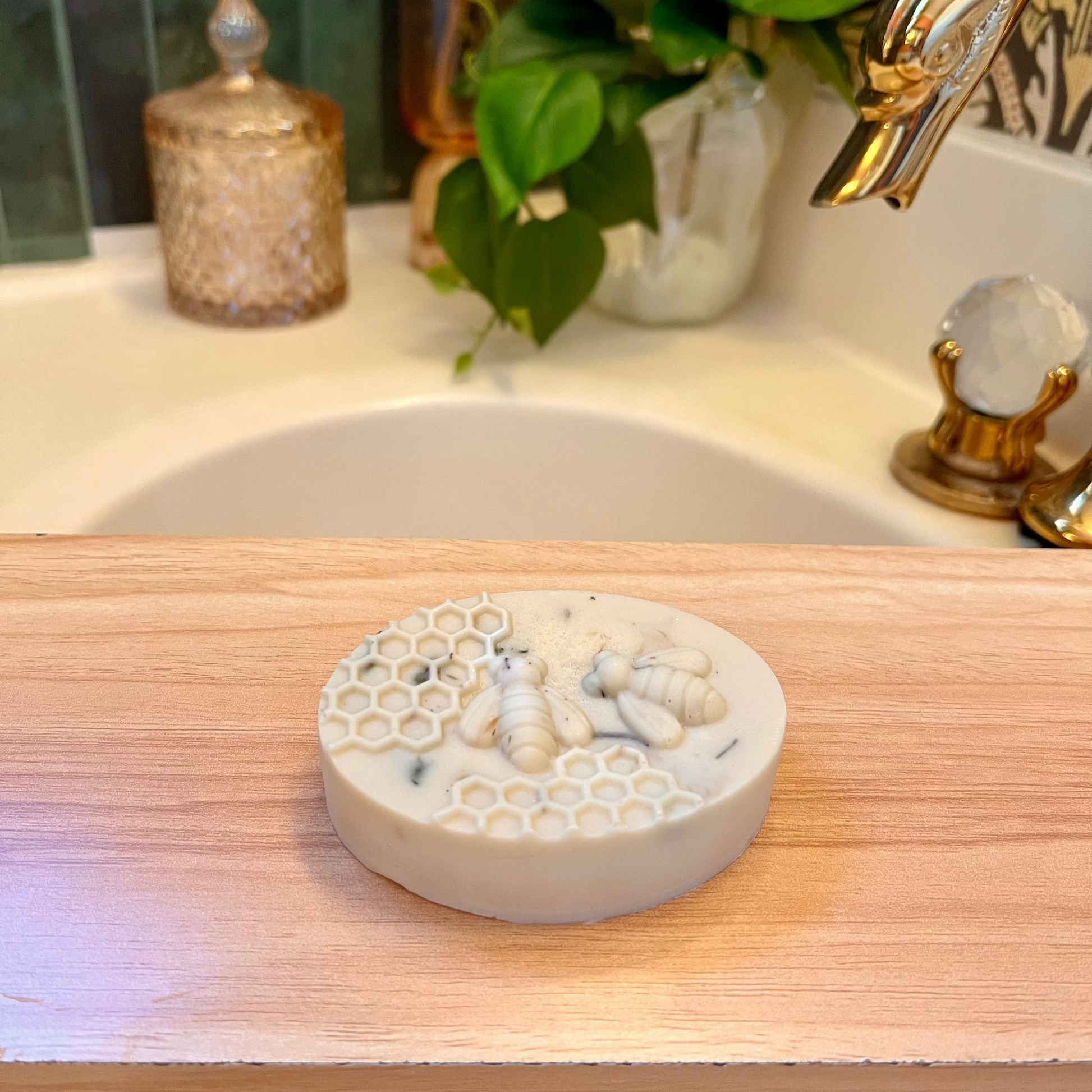 A honey soap bar with a honeycomb pattern, placed on a wooden surface, with a blurred background featuring a sink and a gold-colored faucet.