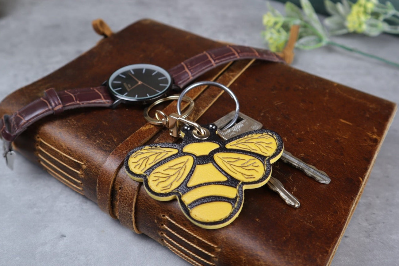 Brown leather notebook with a bee keychain on a gray surface, with a vintage radio and books in the background.